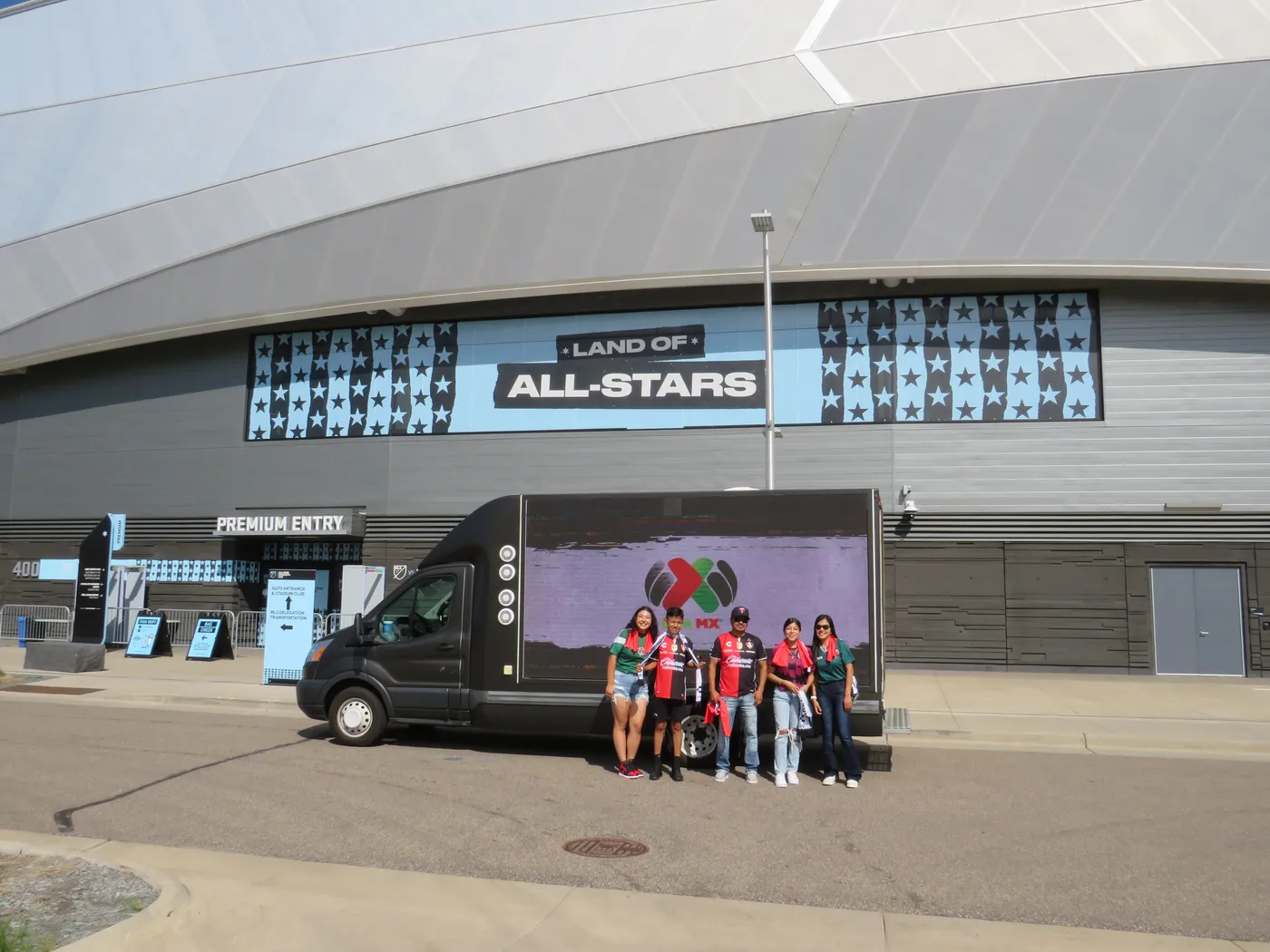 Fans in Liga MX jerseys posing with a Charly digital billboard truck outside Allianz Field under the “Land of All-Stars” banner